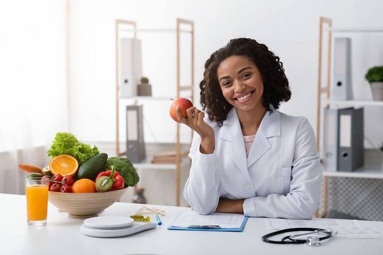 A woman in a medical coat holds an apple, and sits at a table with a large bowl of colorful fruits, a glass of fresh fruit juice, a kitchen scale, a clipboard, and a stethoscope.