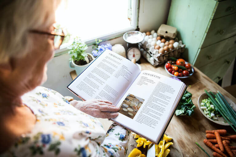 Elderly person holding a cookbook over a table full of fresh eggs and vegetables