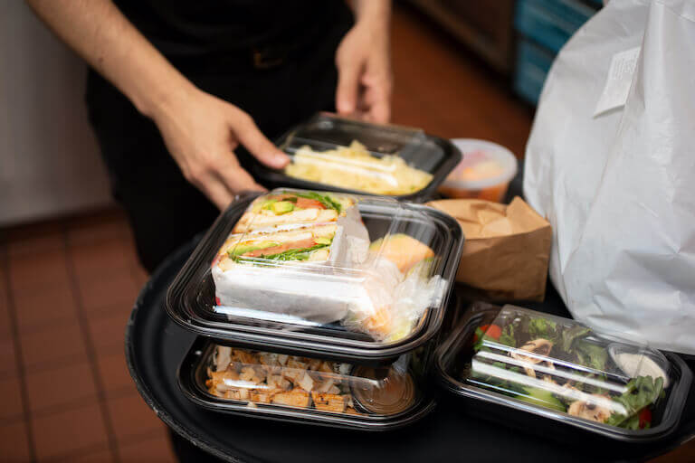 Boxes of to-go foods on a table, with someone closing a lid on a to-go container in the background.