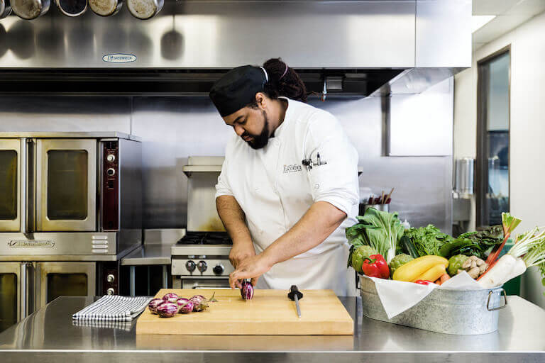 Culinary student in Escoffier uniform slicing vegetables in a teaching kitchen.