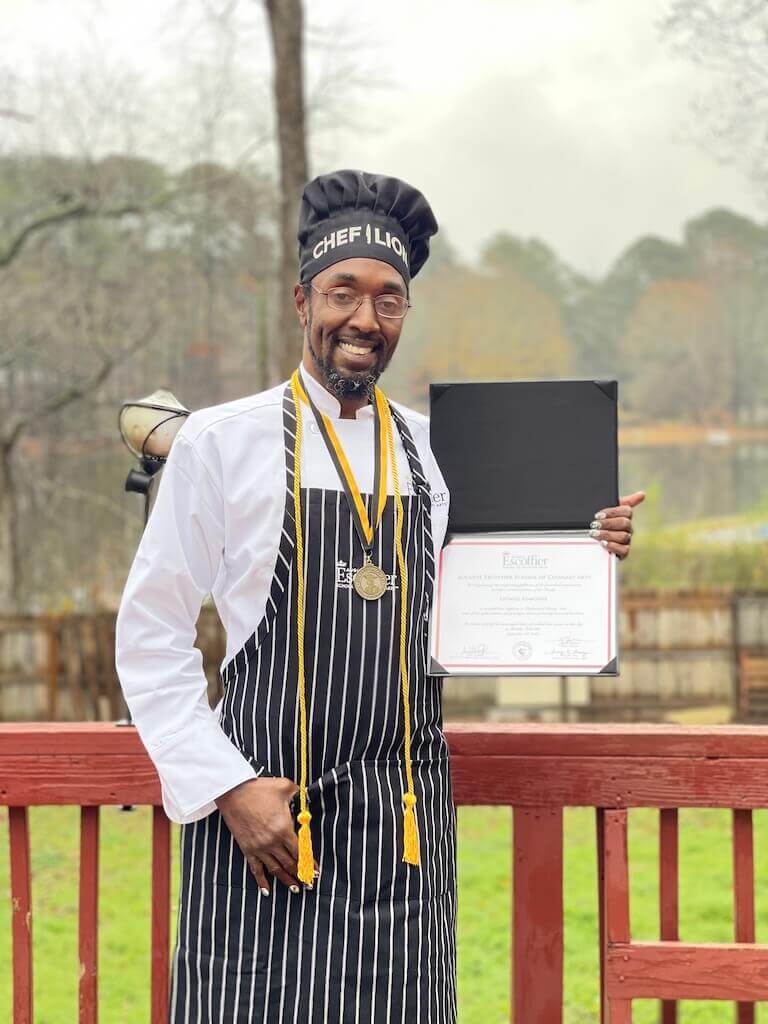 Man in chef uniform and striped apron holding a diploma and medal while smiling outdoors.