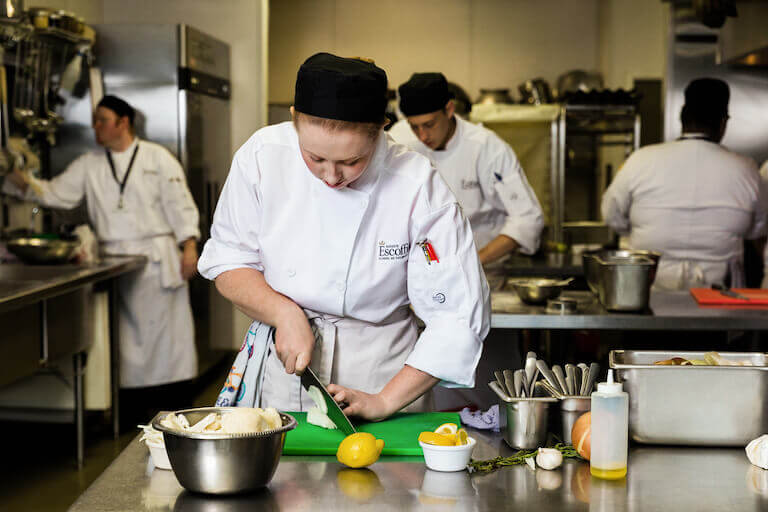 A person in a white chef’s uniform and black skull cap leans over a green cutting board in the foreground of a commercial kitchen.