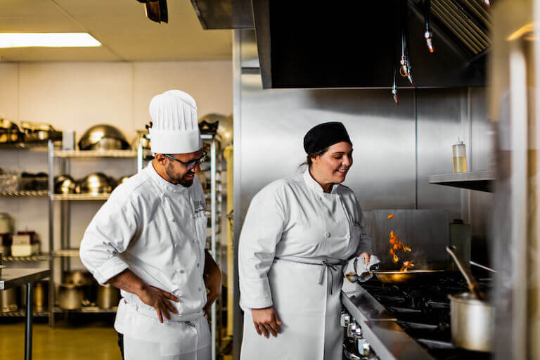 A Chef Instructor looks on while a culinary school student practices sautéeing in a commercial kitchen.