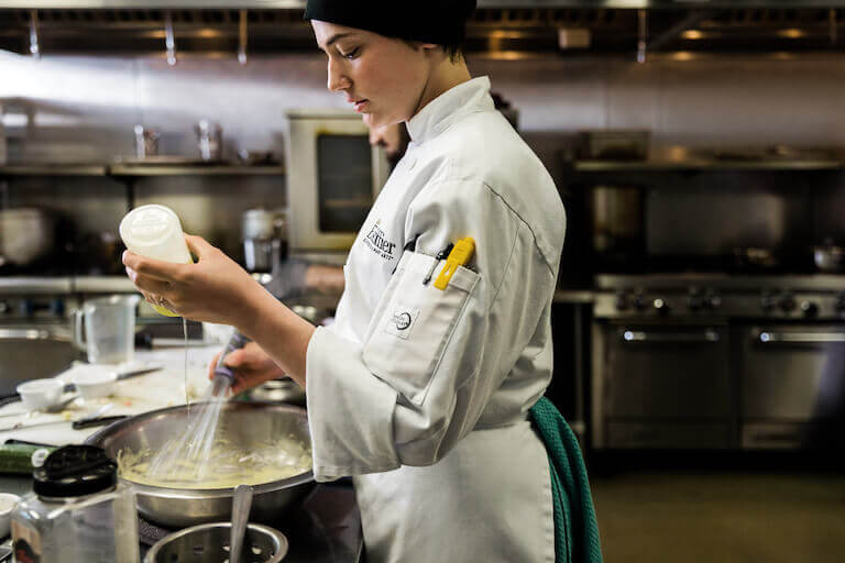 An Escoffier student practices making hollandaise sauce by whisking melted butter into egg yolks in a commercial kitchen.