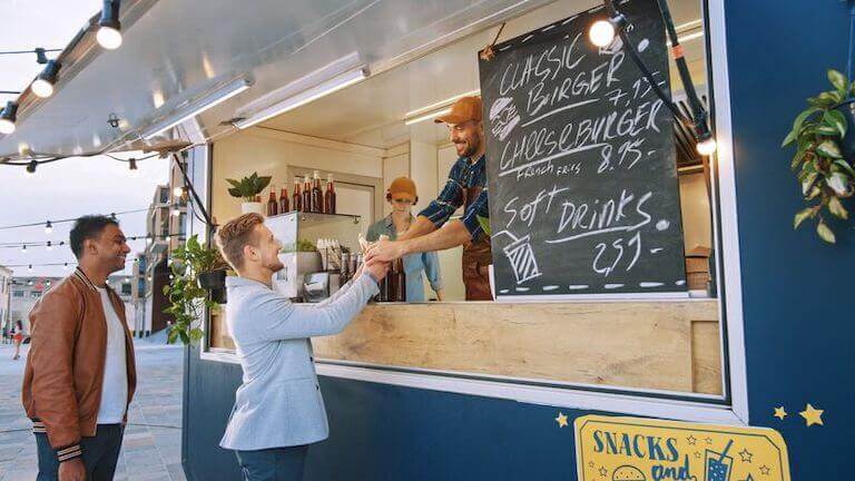 Two food truck customers smiling as a food truck employee hands them their order.