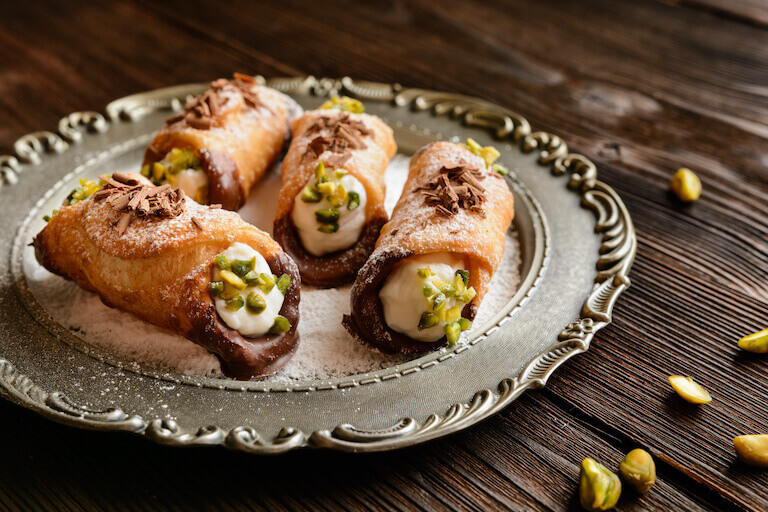 Four cannolis stuffed with ricotta cheese and sprinkled with pistachios sit on a silver plate.