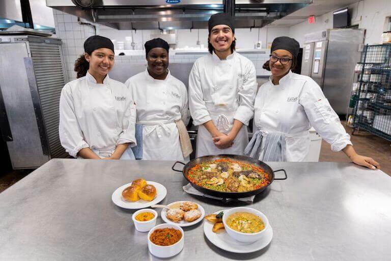 Four culinary students standing behind a counter showcasing their prepared dishes.