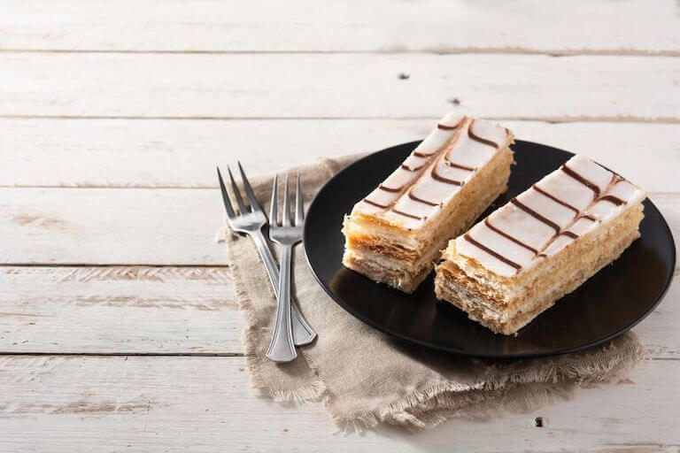Close-up of a neatly layered French mille-feuille pastry with powdered sugar on top, displayed on a white wooden surface.