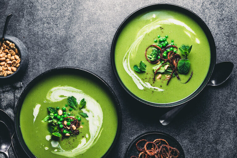 Two bowls of vibrant green pureed soup are artfully displayed with a garnish of green peas, fresh whole mint leaves, caramelized onions, and whole broccoli florets.