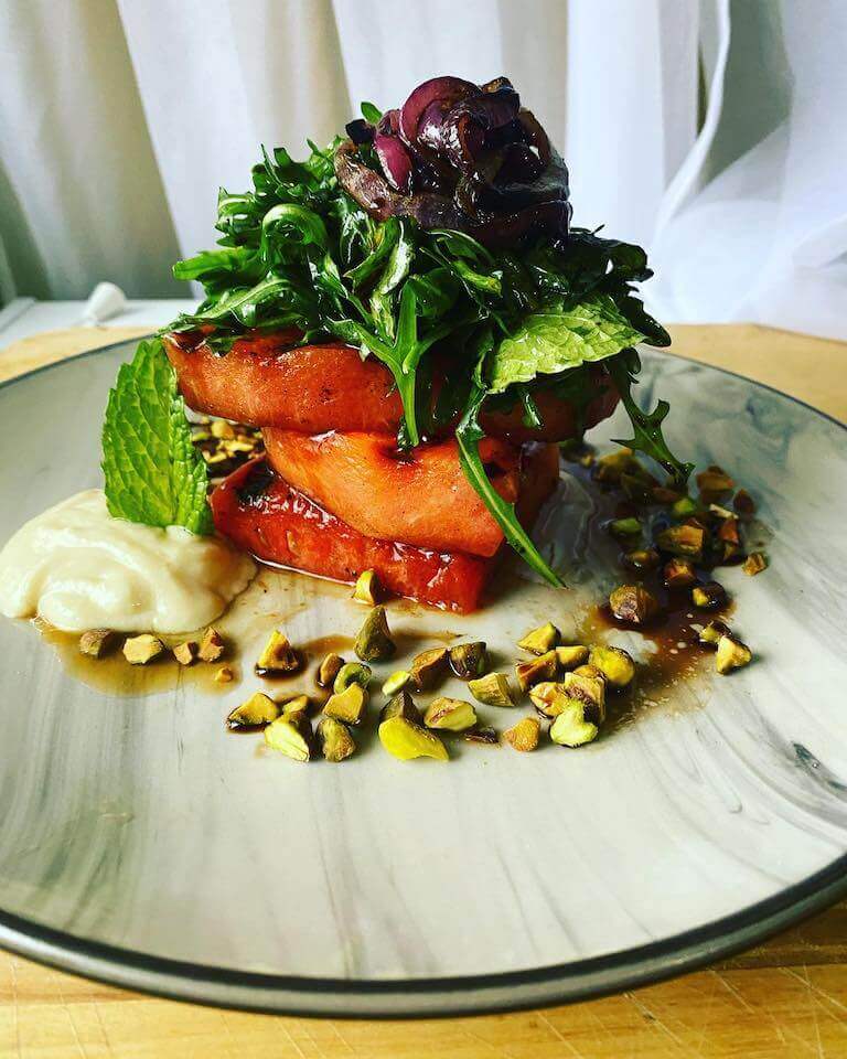 A grilled watermelon salad served on a white and gray plate, garnished with fresh greens and herbs.