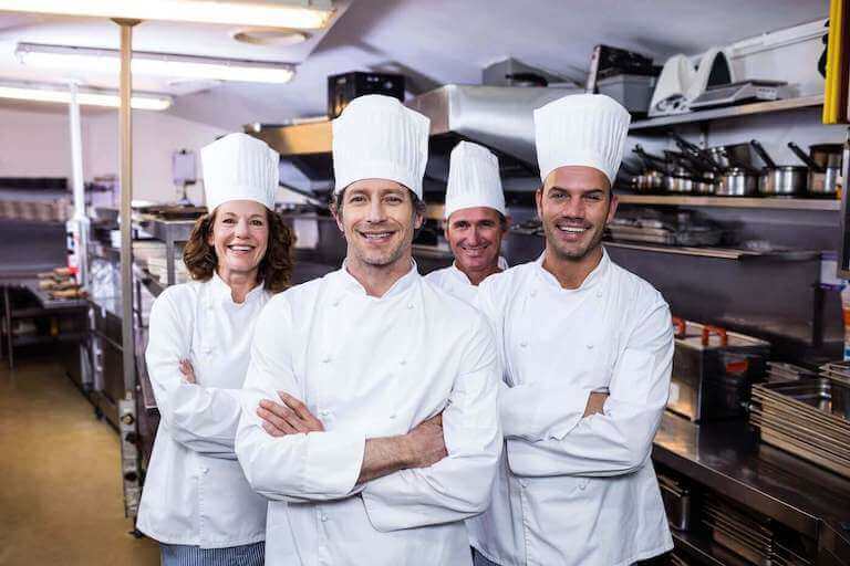 Four smiling chefs standing together in a kitchen wearing white coats and hats.