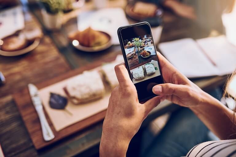 Person using a smartphone to photograph a sandwich on a wooden table for social media.