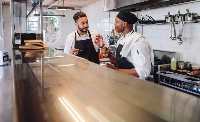 Two chefs in a professional kitchen, both wearing aprons and tasting food with spoons while engaged in conversation.
