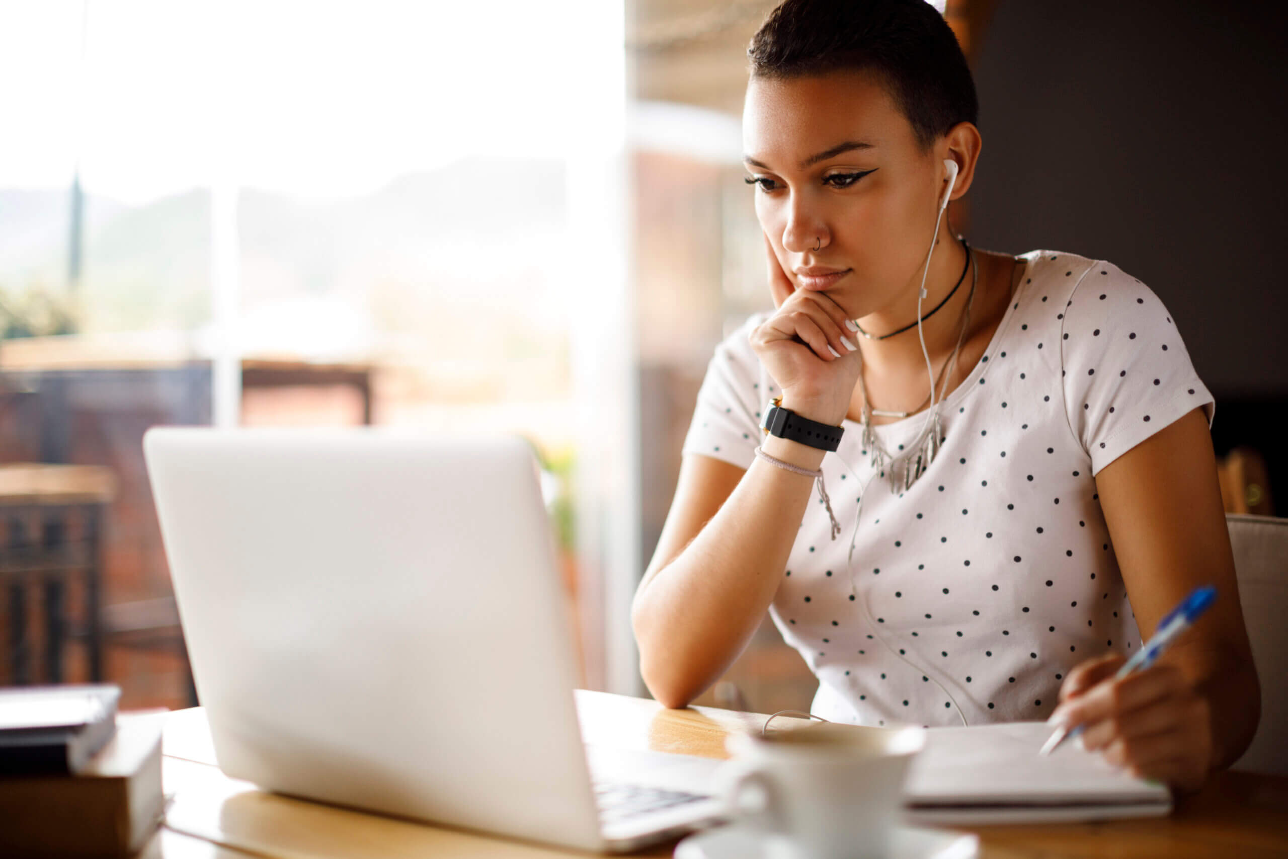 Young woman wearing a white polka dot shirt and earphones looks intently at her laptop while writing notes.