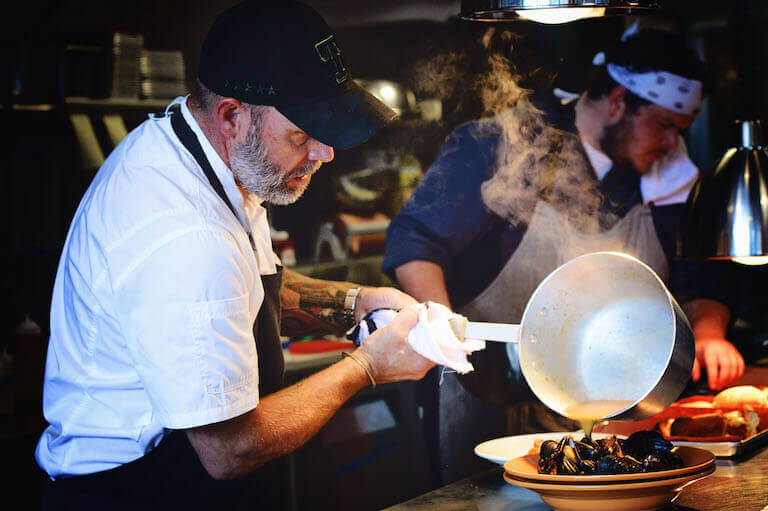 Escoffier graduate Lance McWhorter pouring steaming broth over a bowl of mussels.