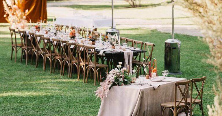 A beautifully arranged outdoor wedding reception with long wooden tables, floral centerpieces, and soft beige tablecloths set on a lush green lawn.