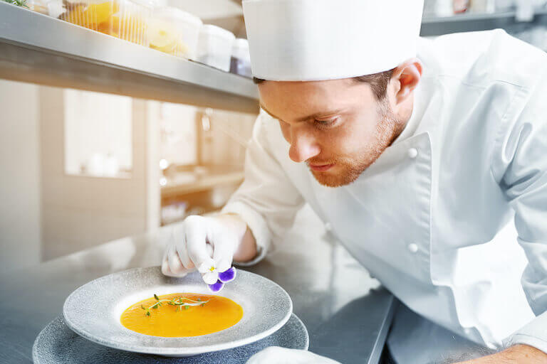 Chef plating and decorating a bowl of soup with a flower at a restaurant kitchen.