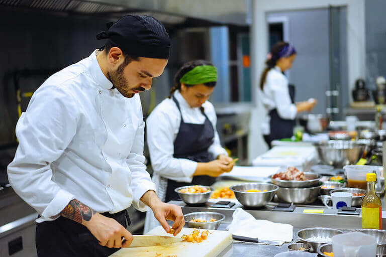 A Commis Chef with a black bandana chopping ingredients on a cutting board in a busy kitchen with other staff working in the background.
