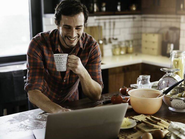 Man holding a mug while using a laptop on the kitchen counter.