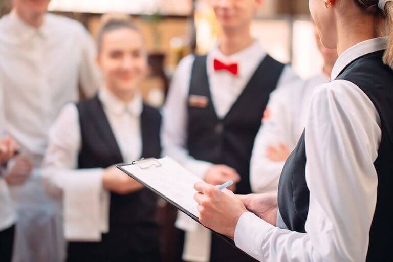 A person holds a clipboard and stands in front of a group of restaurant staff.