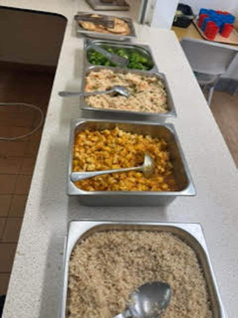 Multiple trays of rice, vegetables, and other prepared dishes laid out on a cafeteria counter.