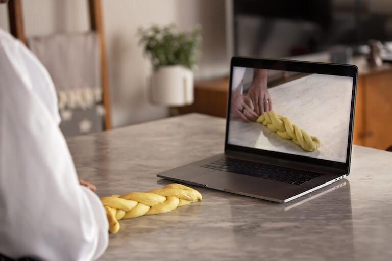 A laptop on a kitchen island shows a pair of hands braiding together dough, while a student mimics the technique on the island.