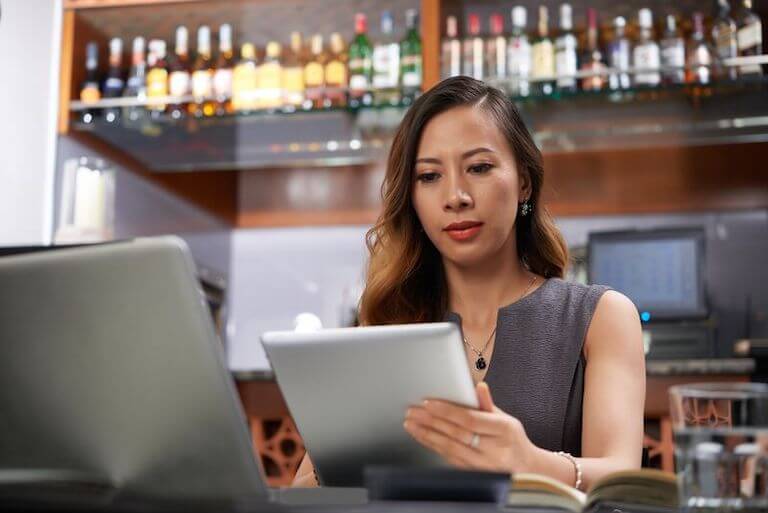 Manager dressed in a grey shirt standing in front of a bar looking at a tablet.