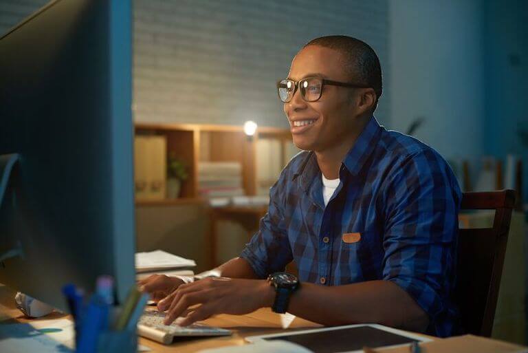 A person in a blue plaid shirt sits at a desk and smiles as they type on a keyboard and look at a large monitor.