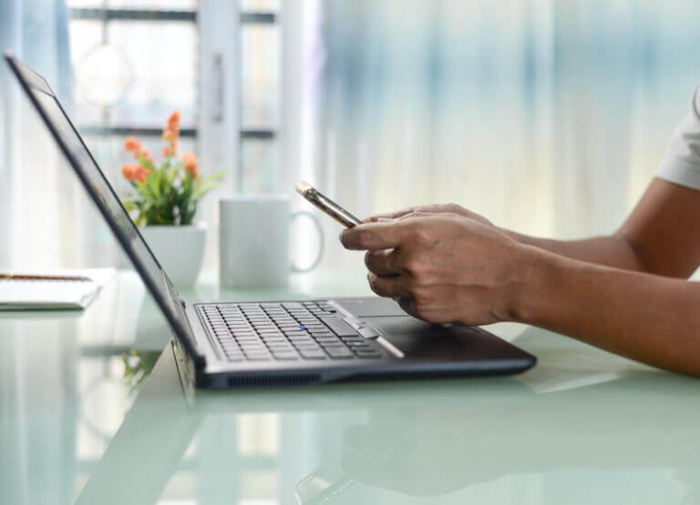 Image shows a closeup of a laptop, and the profile of a person’s arms and hands holding a smartphone.