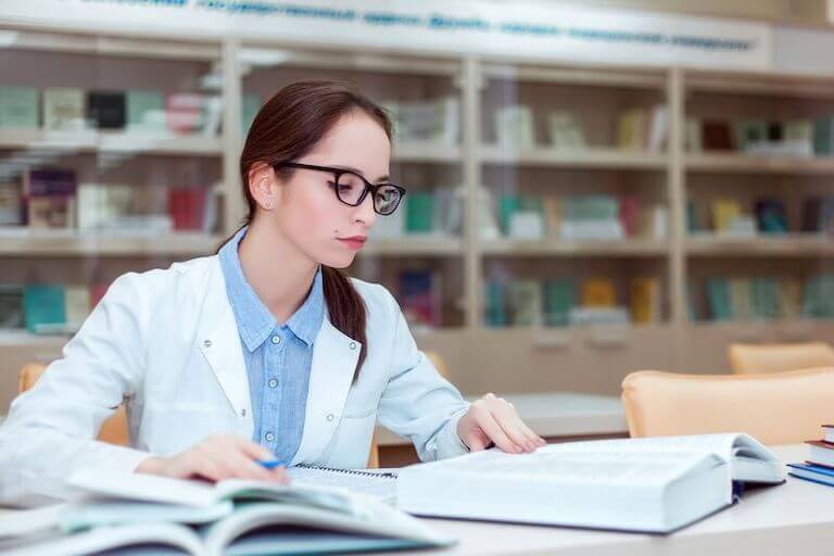 A student in a white medical coat sits at a table studying from multiple textbooks.