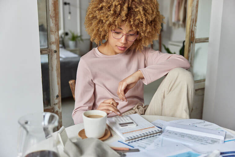 A person with curly hair and glasses sitting at a table cluttered with papers while using a calculator.