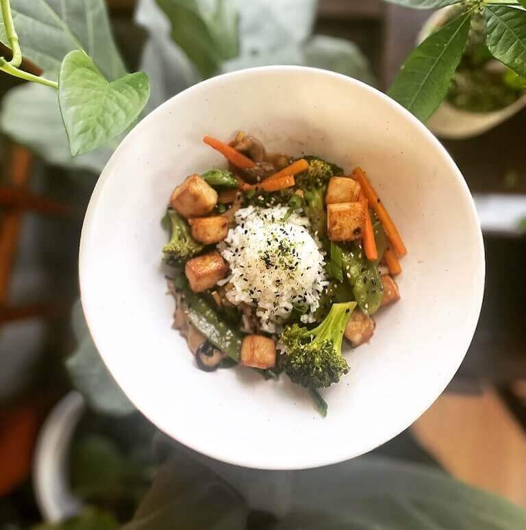 A bowl of tofu stir-fry with broccoli, carrots, snap peas, and rice sits on a table surrounded by green plants, garnished with black sesame seeds and seasoning.