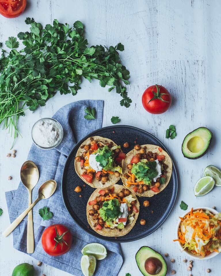 A plate with three small tacos topped with beans, lettuce, guacamole, tomatoes, and lime, surrounded by fresh vegetables, like cilantro, avocados, and whole tomatoes.