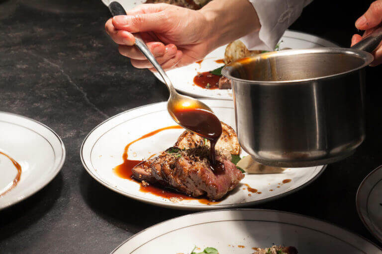 A chef’s hand pours a rich, dark sauce over a plate of sliced beef.