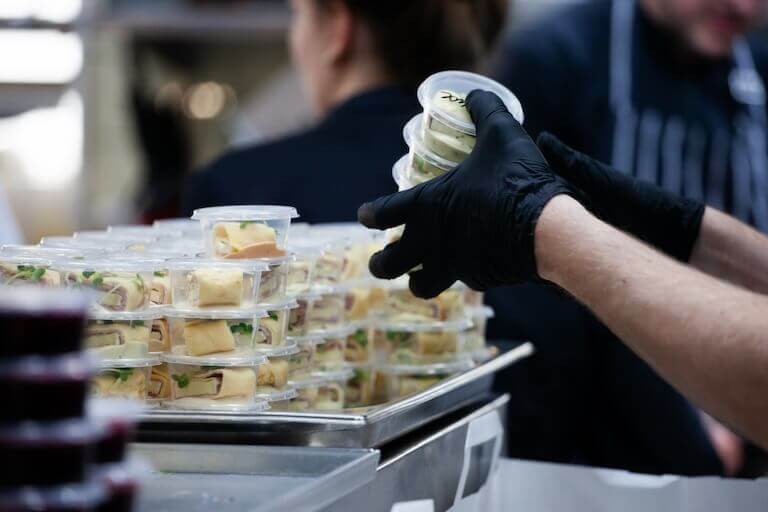 A person wearing latex gloves picks up several appetizers from a tray of individually-packaged appetizers in a professional kitchen setting.
