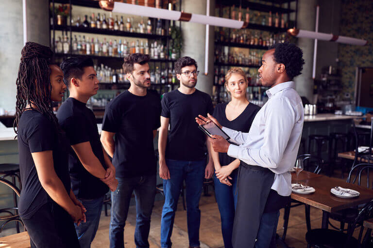 A restaurant manager speaks to a group of five employees in a restaurant that has not yet opened.