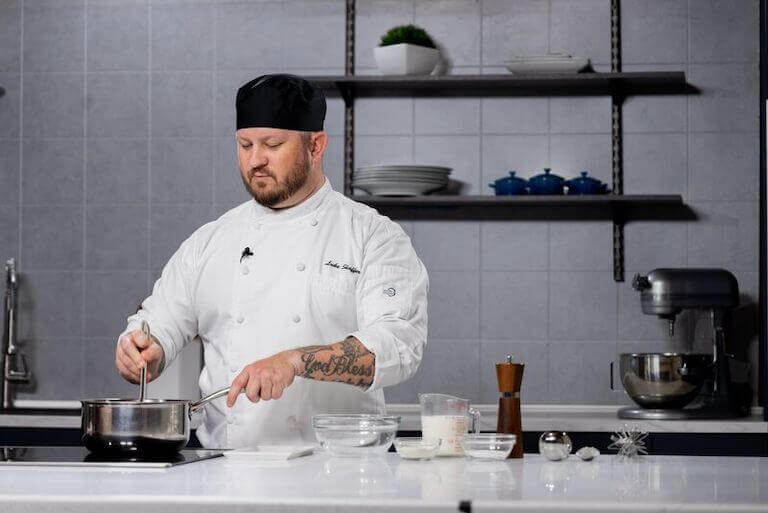 Saucier stirring gravy in a saucier pan with measured ingredients in glass bowls next to the stove.