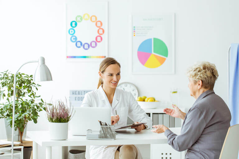 A woman in a white coat sits across a desk from a coaching client, who is discussing her health challenges.