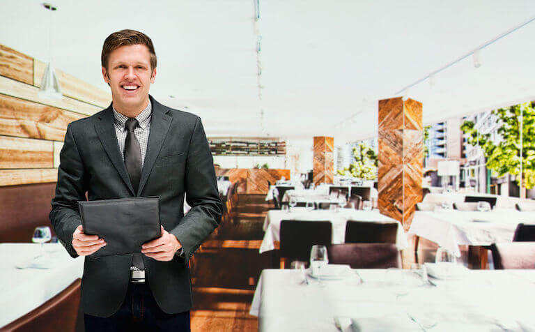 A person wearing a suit and tie stands in the foreground of a restaurant, facing the camera and smiling.
