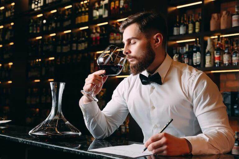 A sommelier in a white shirt and black bow tie swirls a glass of red wine, analyzing its aroma while making tasting notes at a dimly lit bar.