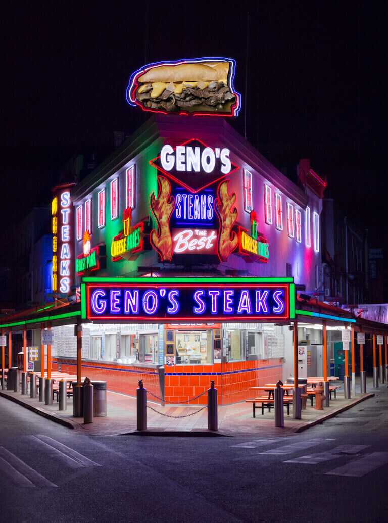 The front facade of Geno’s Steaks is on a triangular corner and is lit up with many differently colored neon signs, some of which read “Geno’s Steaks”, and “Cheese Fries.”