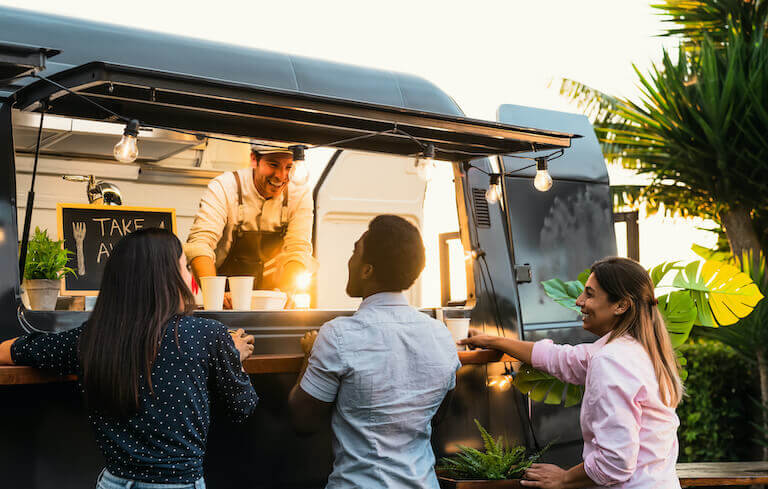 A chef in an apron stands in a food truck talking with several smiling customers surrounded by tropical foliage.