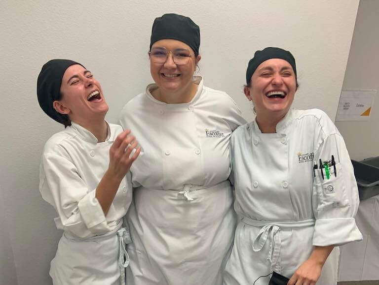 Three Escoffier culinary students wearing white chefs coats and laughing together.