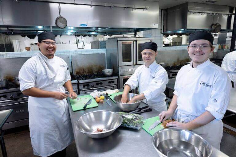 Three Escoffier students mix salads in large stainless steel bowls in a commercial kitchen. They are wearing Escoffier uniforms and smiling at the camera.