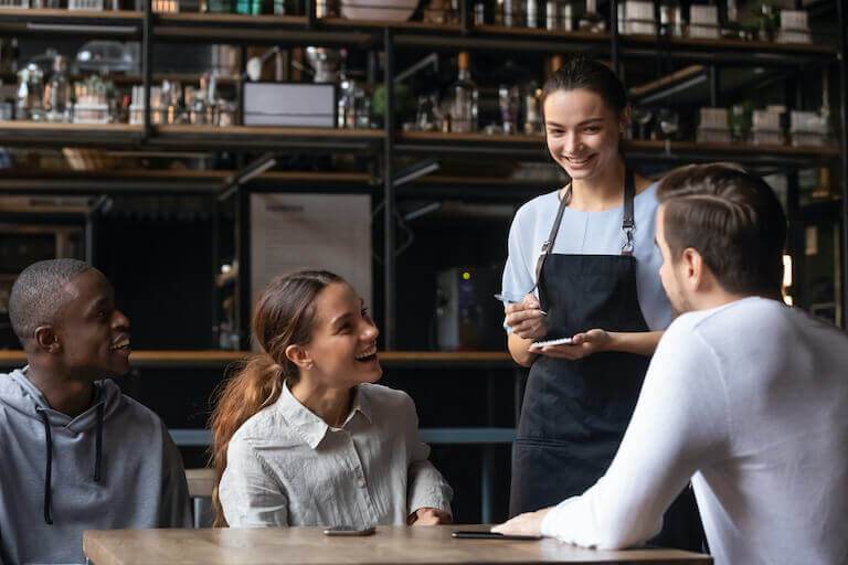 A restaurant server with pad and pen stands at a table of three guests and smiles. The guests are smiling and laughing.