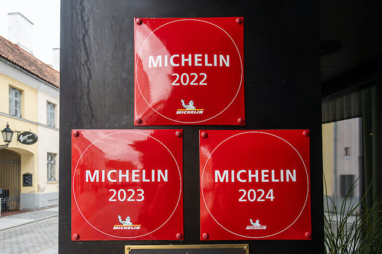 Three square red signs read "Michelin 2022" "Michelin 2023" and "Michelin 2024" on a restaurant door frame.