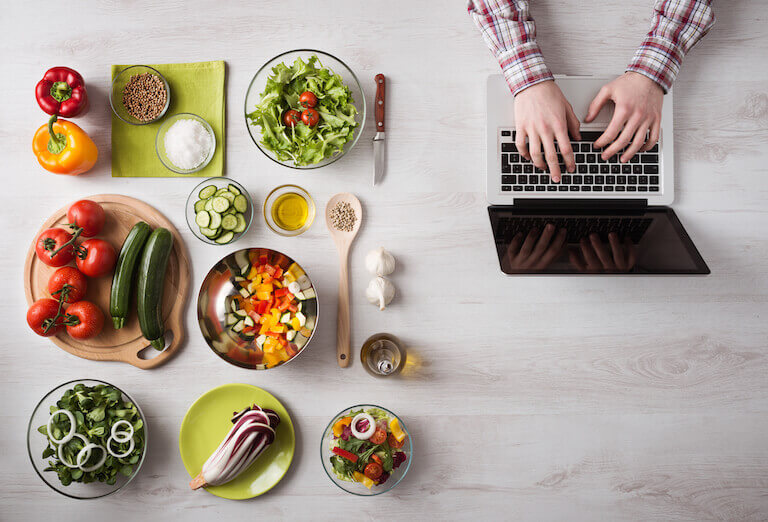 Over-head shot of hands typing on a laptop, next to mise-en-place of various fresh produce, spices, and cooking utensils on a wooden table.
