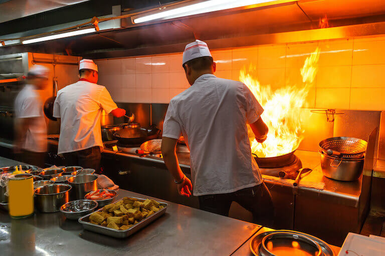 Two chefs stirring woks with a large flame