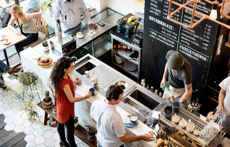Several people stand at the counter of a busy cafe, while two employees work behind the counter preparing drinks.