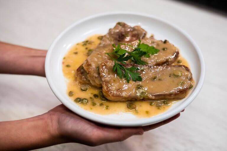 Two hands holding a dish of seitan piccata, served on a white plate with a garnish.